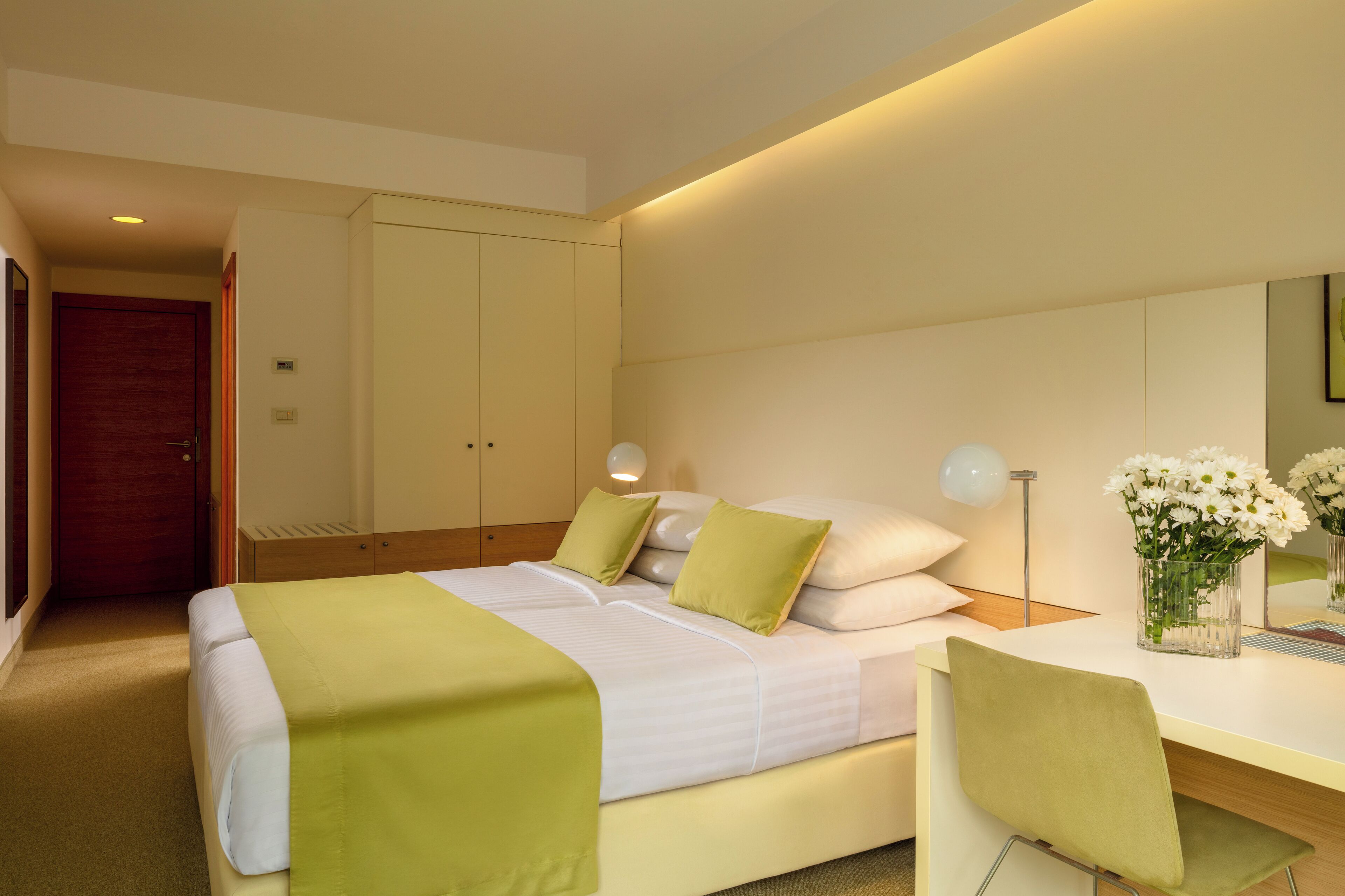 A hotel room with a bed, a desk with a chair, a vase of flowers, and a door.