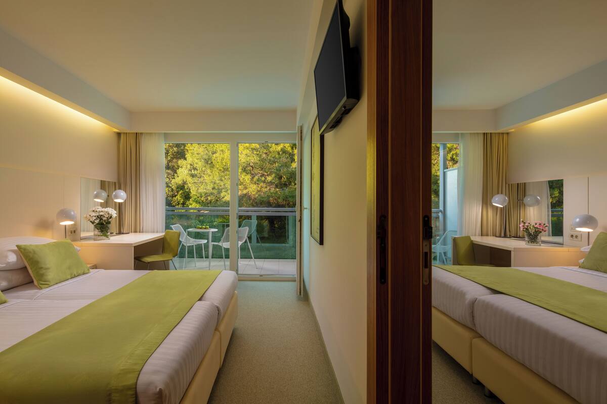 A hotel room with two beds, a desk, a chair, a TV, and a balcony with a view of greenery.