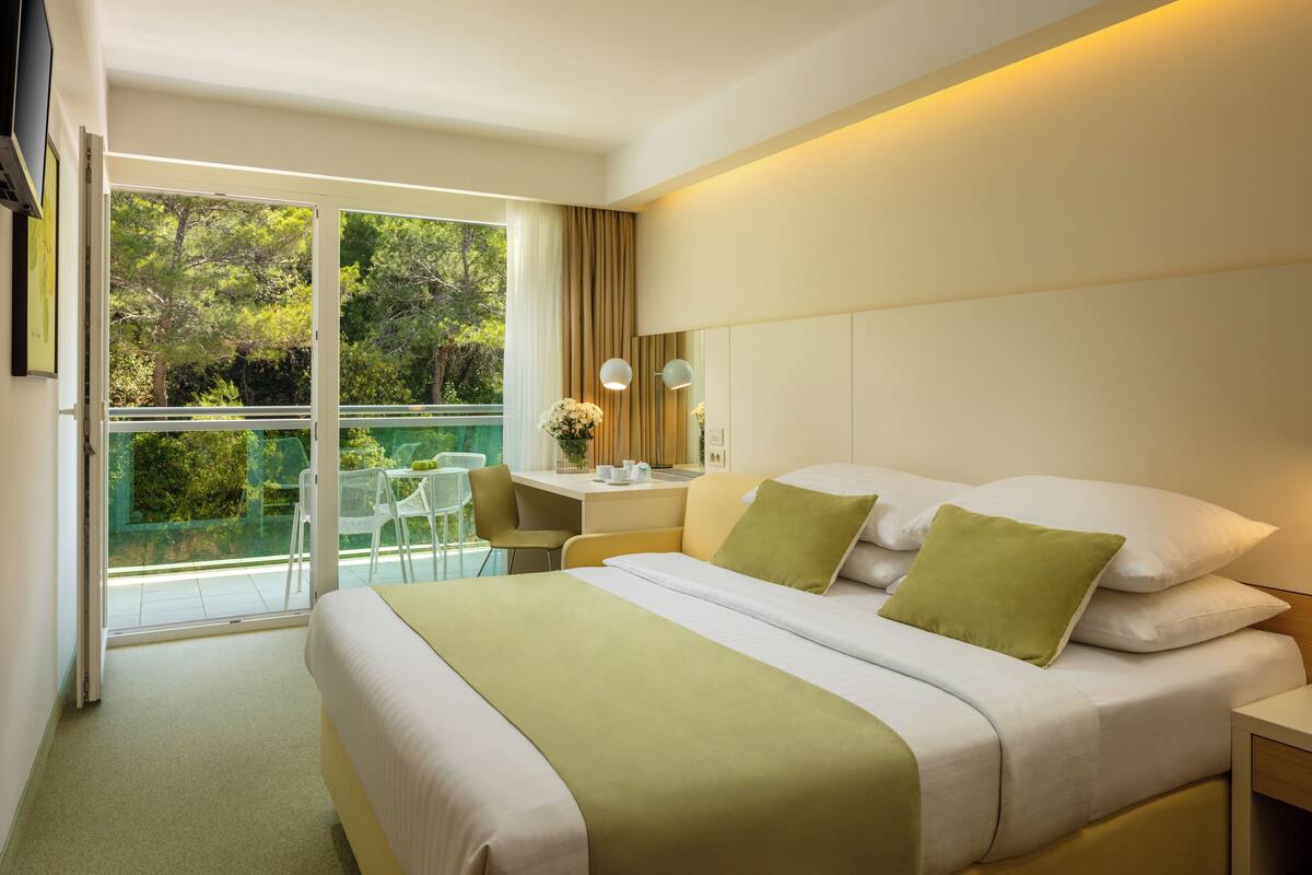 A modern hotel room with a large bed, a desk, and a balcony with a view of trees.