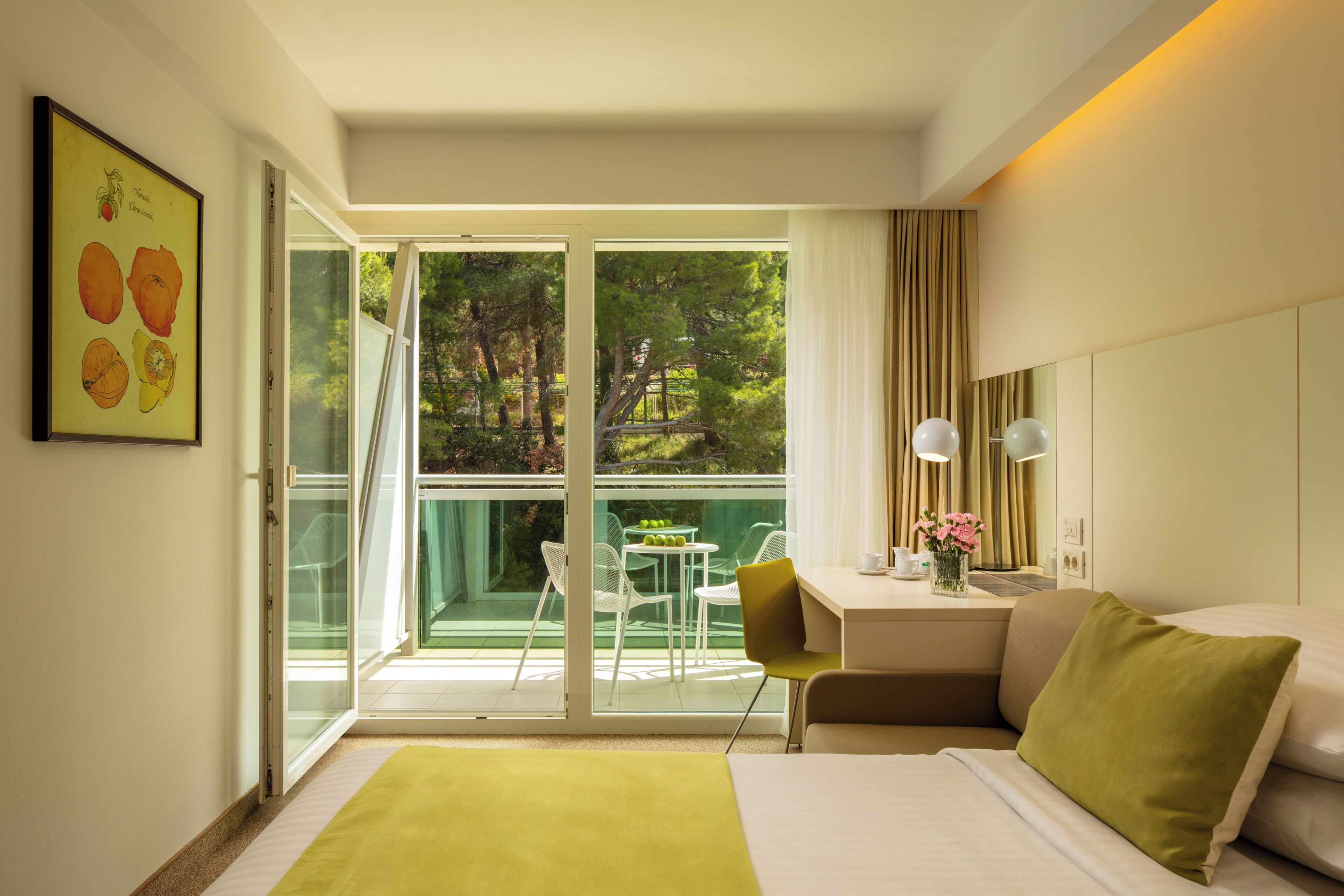 A hotel room with a bed, a desk, a chair, a table with a vase of flowers, and a balcony with a table and chairs.