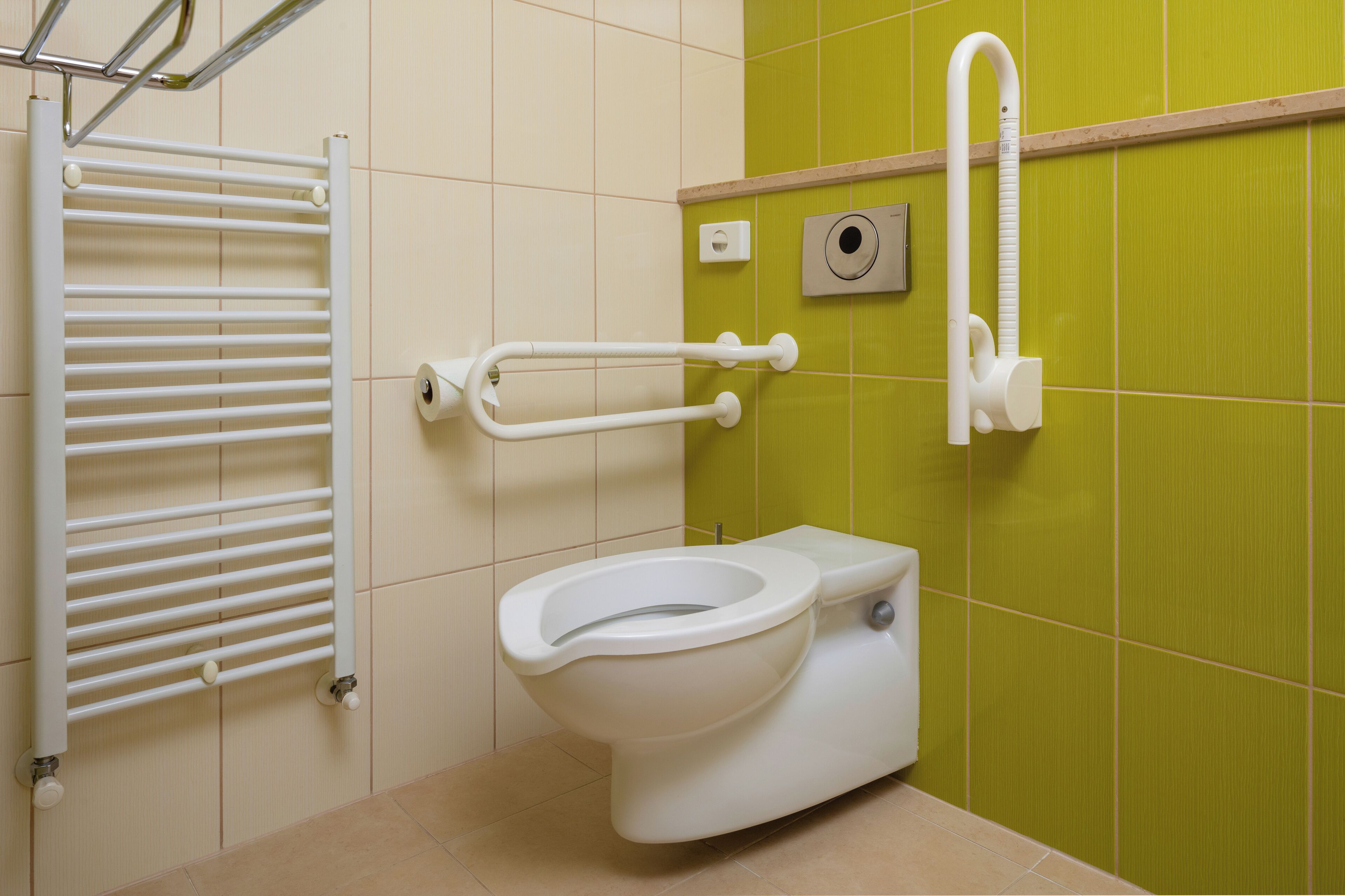 Une salle de bain avec des toilettes blanches, une barre d’appui, un porte-serviettes et un mur recouvert de carreaux verts.