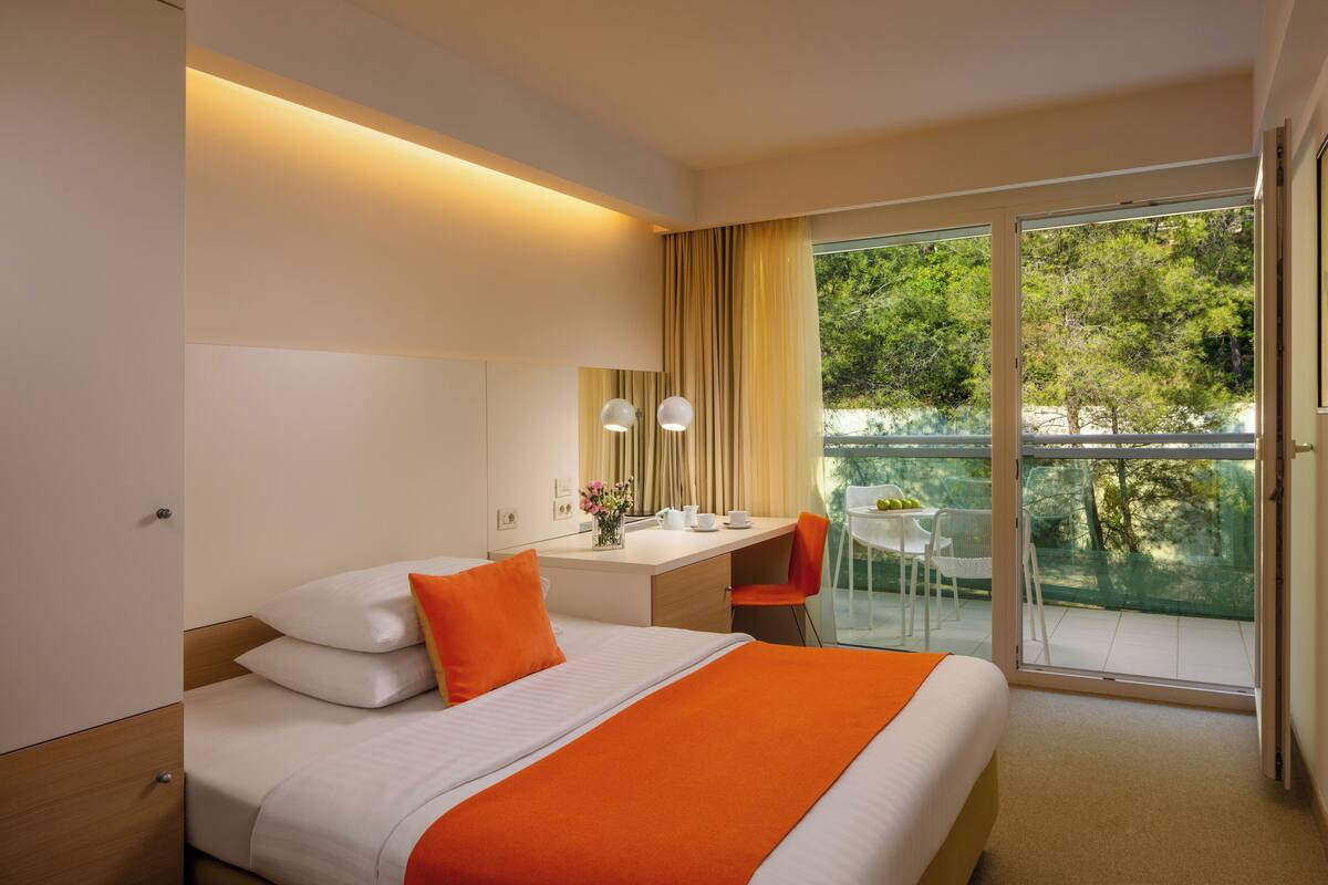 A hotel room with a bed, a desk, a chair, a small table with a vase of flowers, and a balcony with a glass railing.