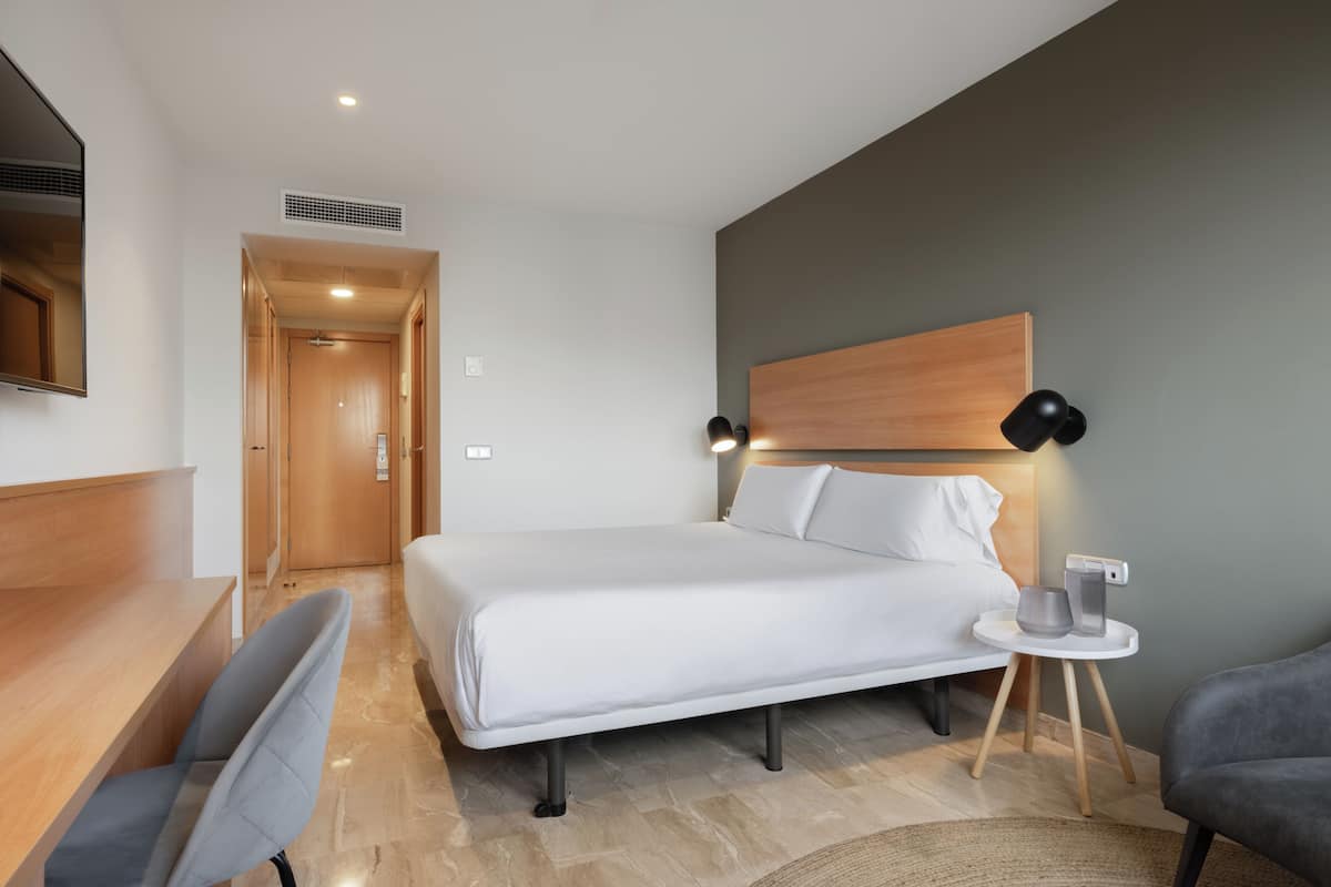 Ein modernes Hotelzimmer mit Bett, Schreibtisch, Stuhl und einem kleinen Tisch mit Lampe.