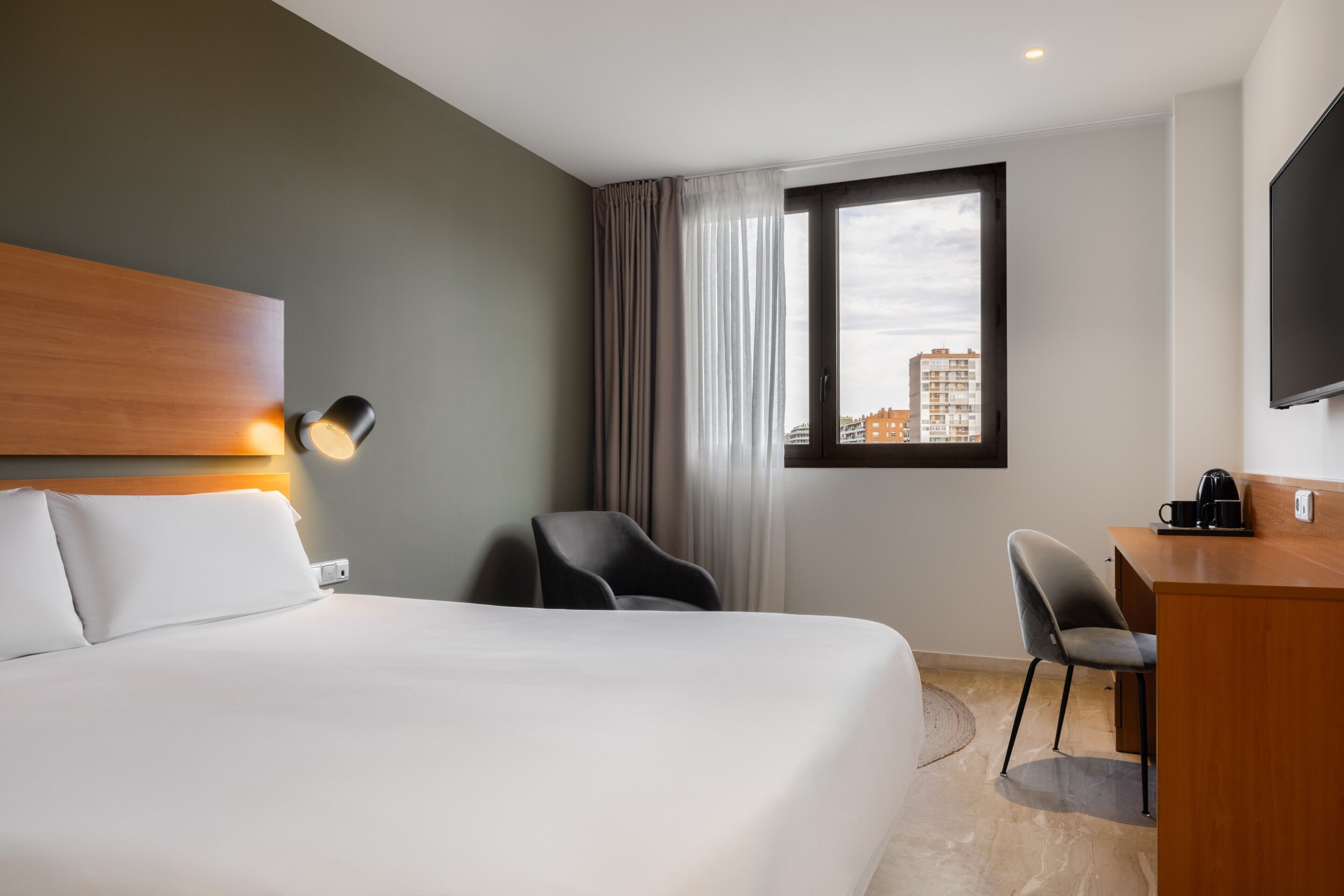A hotel room with a bed, a desk, a chair, a lamp, and a view of buildings.