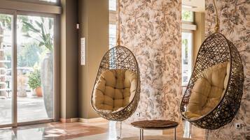 Two hanging chairs with cushioned seats and a woven frame, positioned on a round wooden table in a room with patterned wallpaper and large windows.