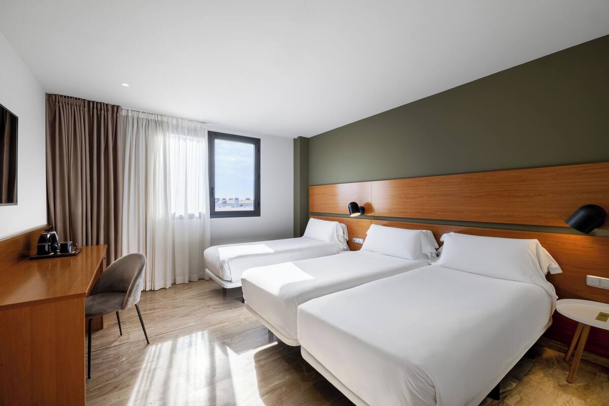 Ein Hotelzimmer mit zwei Betten, einem Schreibtisch mit Stuhl, einem kleinen Tisch und einem Fenster mit Vorhängen.