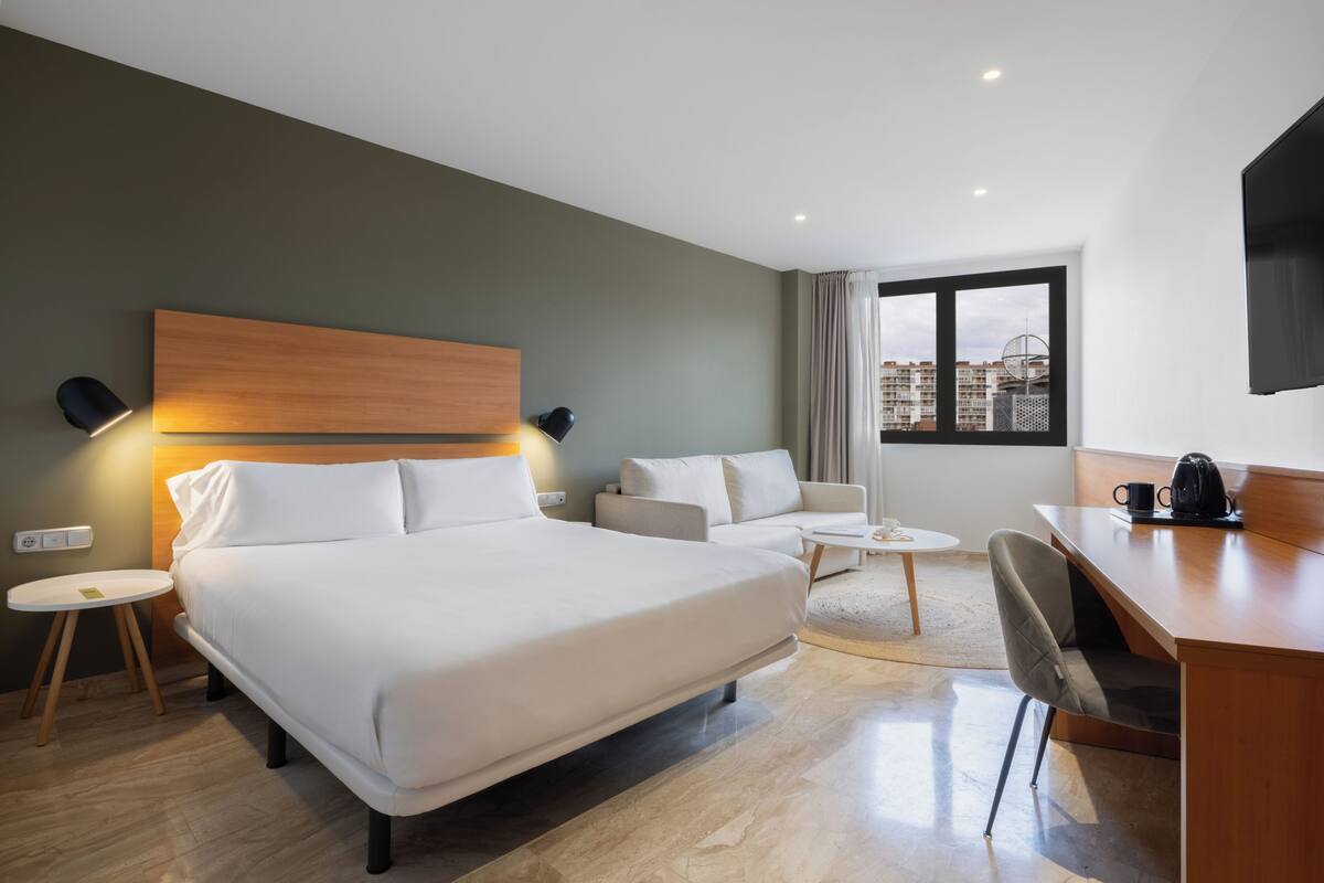 Ein Hotelzimmer mit Bett, Schreibtisch, Stuhl, rundem Tisch und Blick auf die Stadt durch ein Fenster.