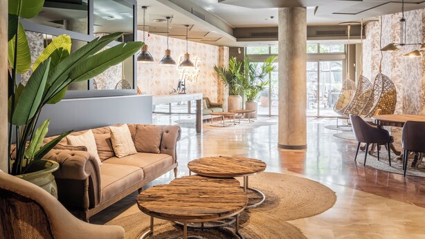 Un vestíbulo amplio con un sofá, dos mesas redondas de madera, luces colgantes y un cartel que dice 'to travel'.