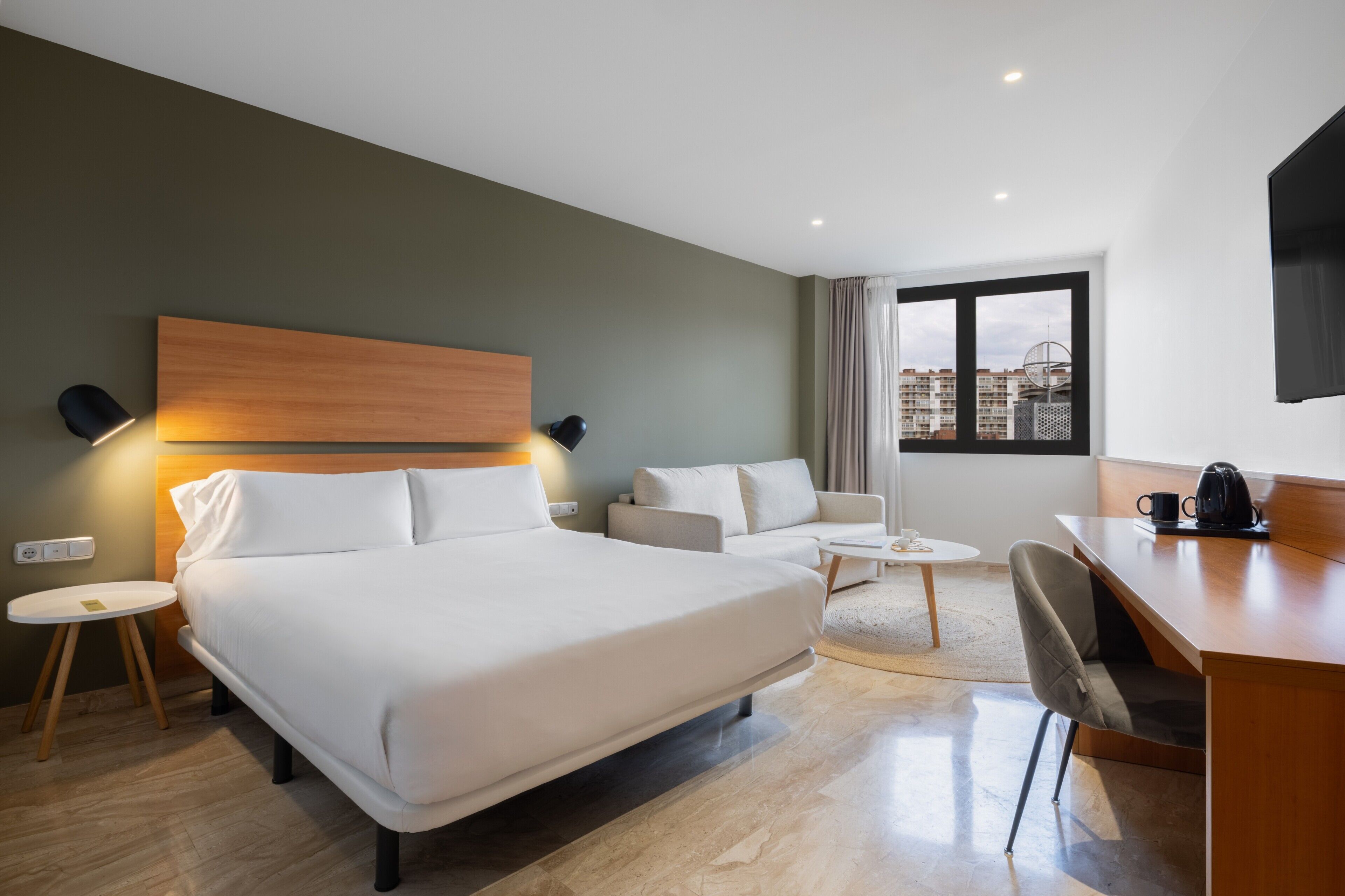 A modern hotel room with a large bed, a sofa, a desk, and a view of the city.