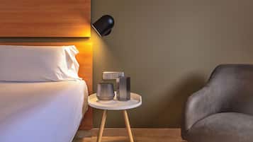 A neatly made bed with white linens, a small bedside table with two metallic vases, a modern lamp, and a gray upholstered chair.