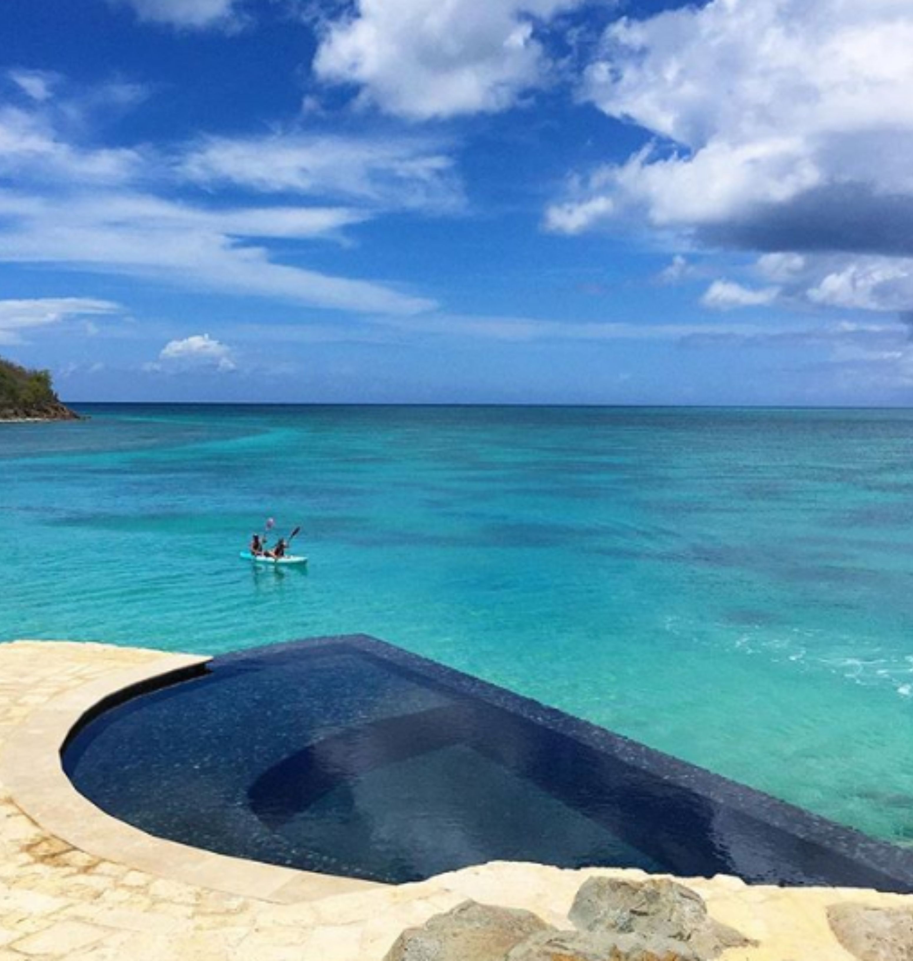 A clear blue sea with a small boat and a stone infinity pool.
