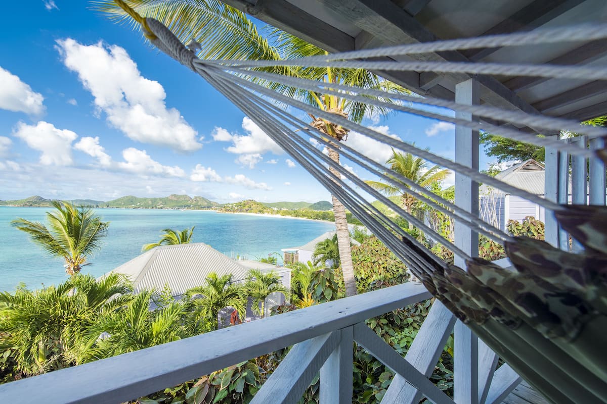 A hammock view overlooking a turquoise sea, palm trees, and a white building with a sloped roof.