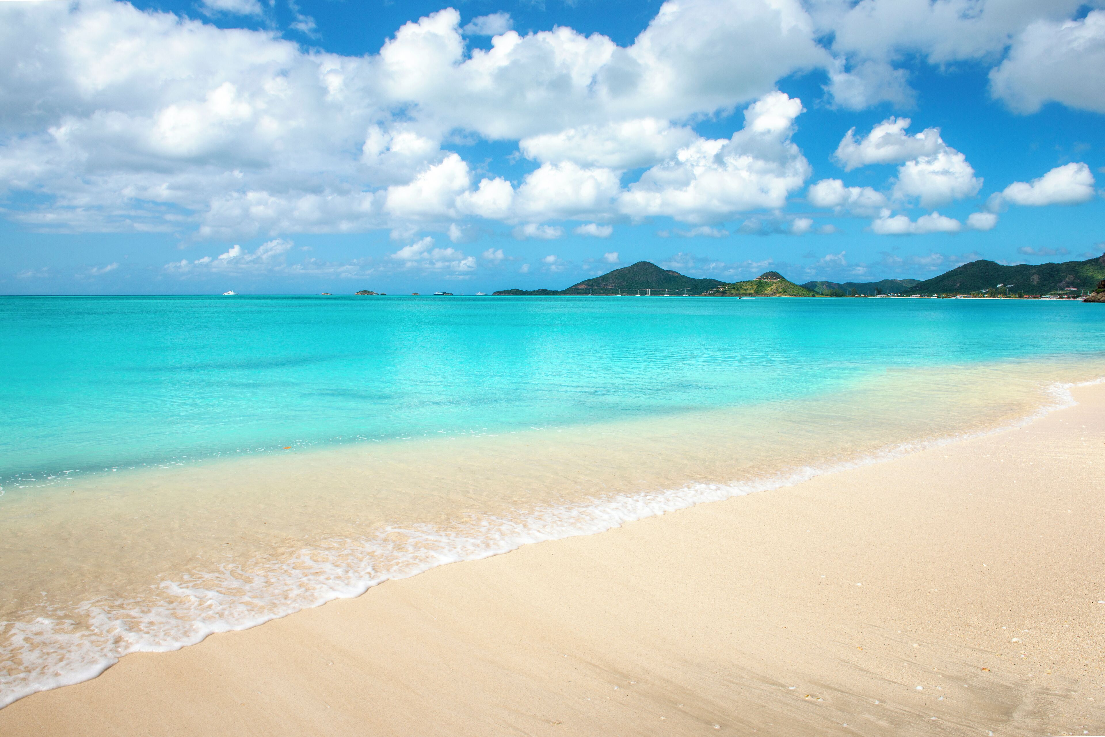 A beach with clear turquoise water, gentle waves, and distant green hills under a partly cloudy sky.