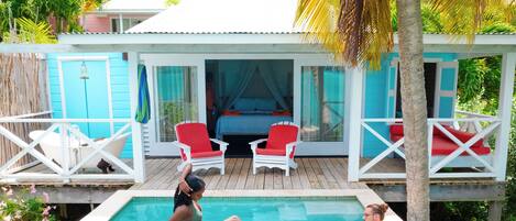 A pool area with a wooden deck, two red chairs, and a glass-enclosed bedroom with a bed and curtains.