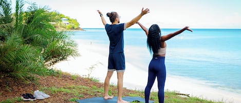 Two people on yoga mats at a beach with a clear view of the ocean and a house in the background.