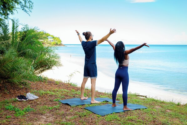 Two people on yoga mats at a beach with a clear view of the ocean and a house in the background.