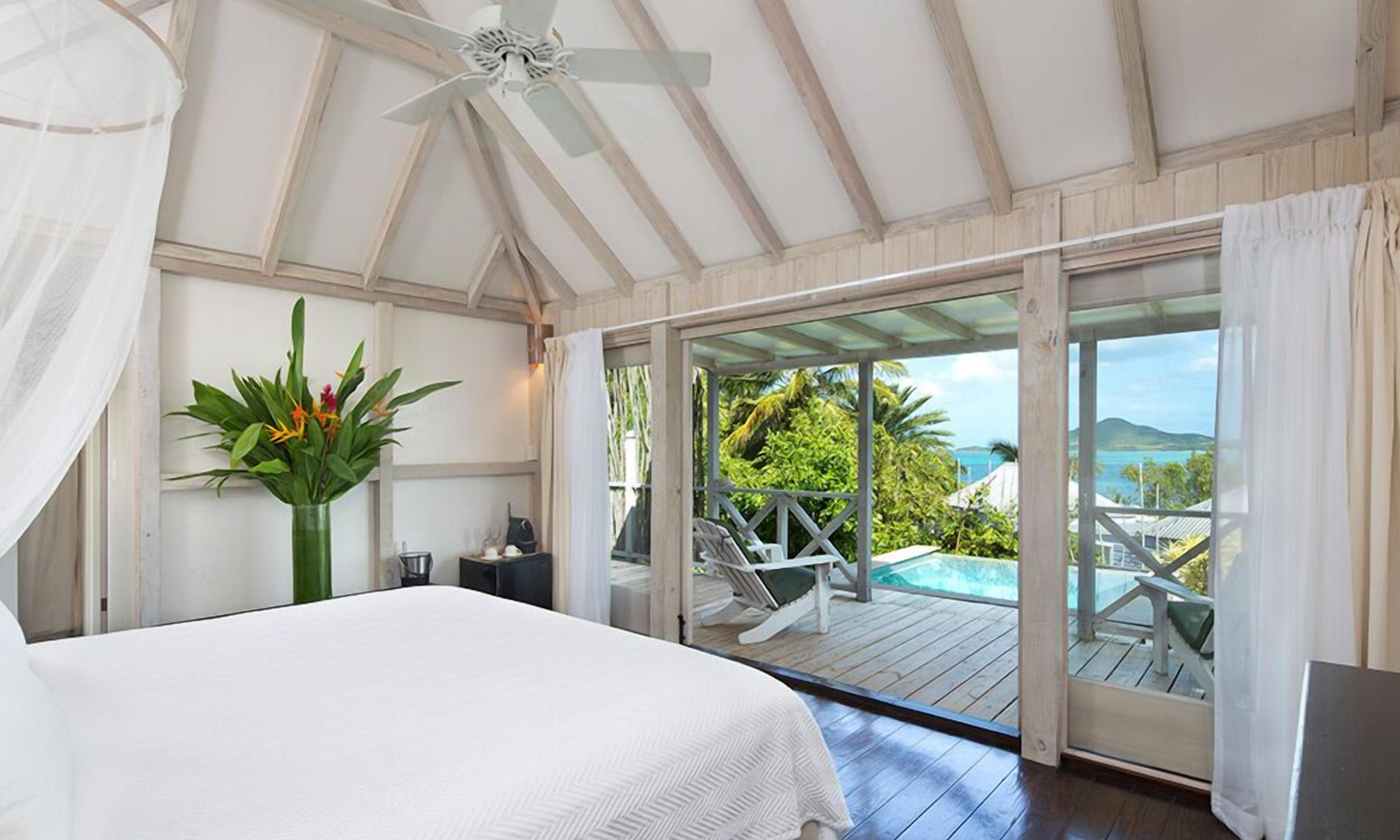 A bedroom with a bed, a ceiling fan, a large window, and a view of a pool and greenery.