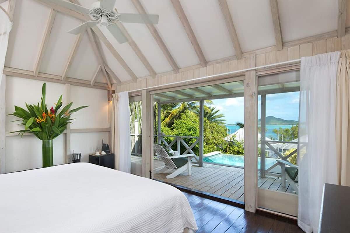 A bedroom with a bed, a ceiling fan, a large window, and a view of a pool and greenery.
