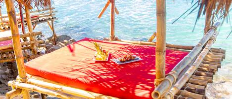 Une chaise de plage en bambou, dotée d’un coussin rouge et accompagnée d’une assiette de nourriture, et offrant une vue sur la mer.