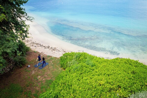 A beach with clear water, two people on a mat, and greenery.