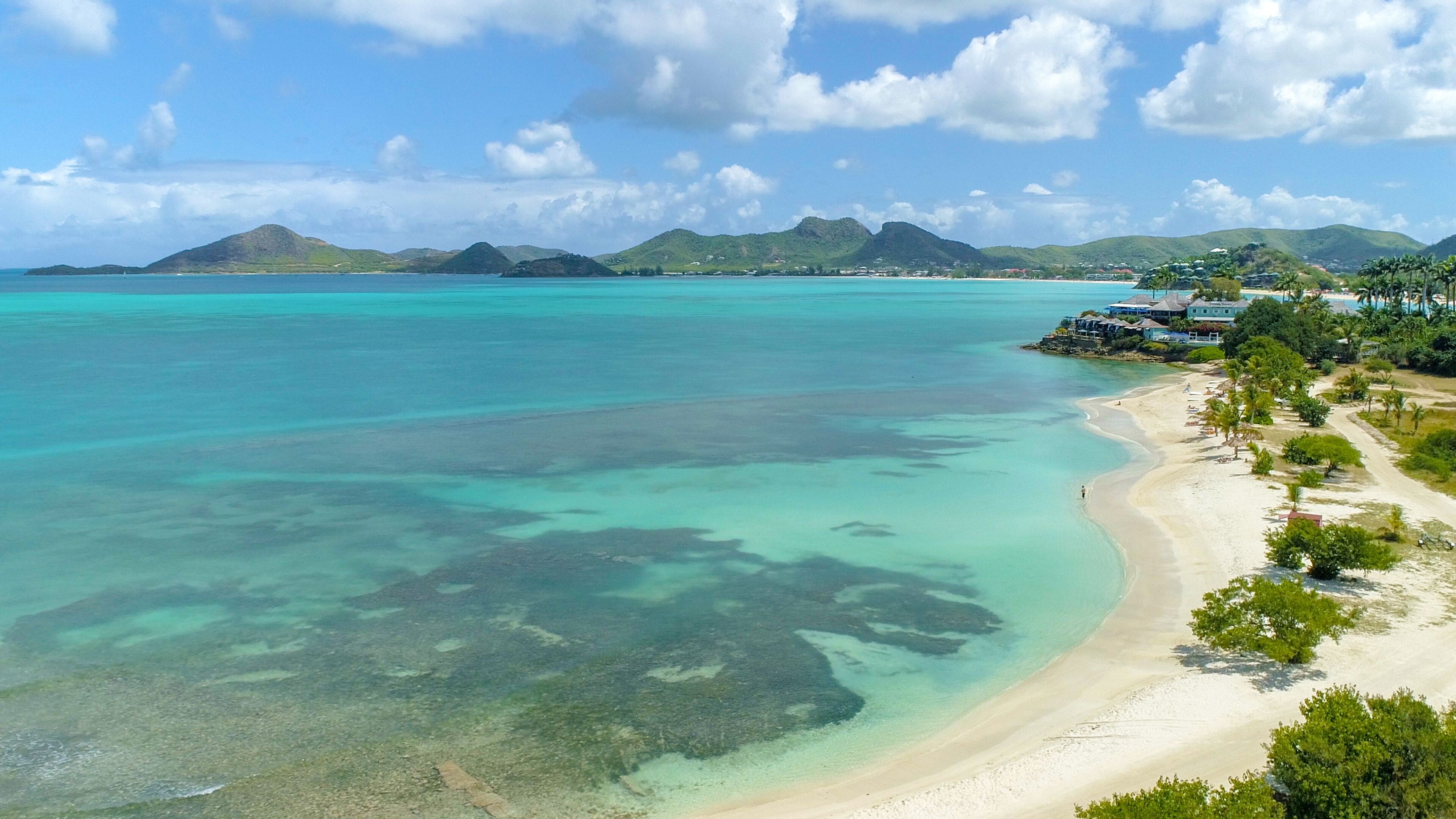 A beach resort with clear turquoise waters, a sandy shoreline, and a backdrop of green hills.