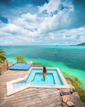 A wooden deck with a swimming pool, a person in a swimsuit, and a view of the ocean with a sailboat in the distance.
