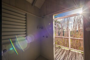 A room with a wooden ceiling, a window with blinds, and a view of a wooden deck and palm trees outside.