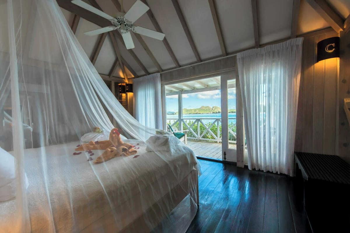 A bedroom with a canopy bed, a ceiling fan, a view of the sea, and a balcony with a railing.