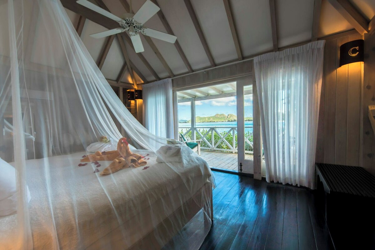 A bedroom with a canopy bed, a ceiling fan, a view of the sea, and a balcony with a railing.