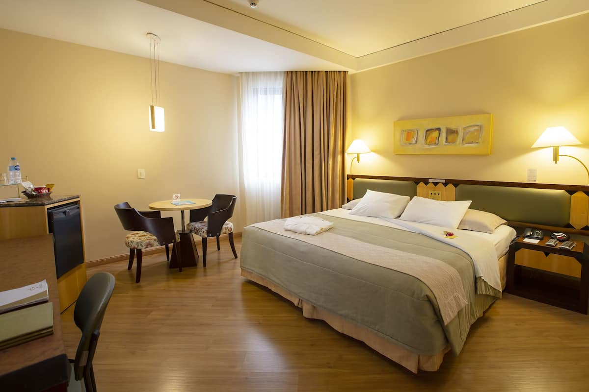 A hotel room with a bed, bedside tables, a desk with chairs, a small table, and a window with curtains.