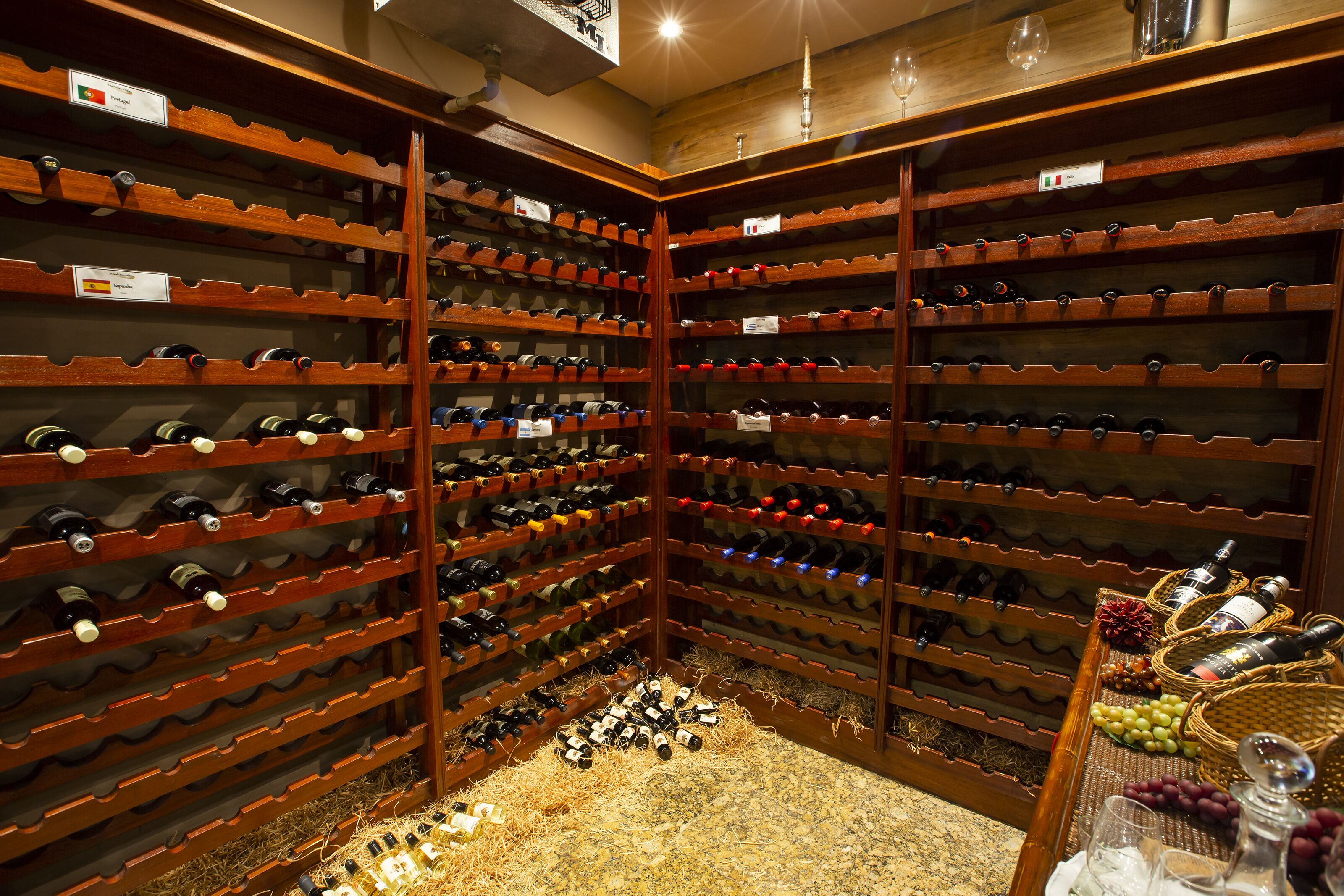 Una bodega con múltiples estantes de madera llenos de botellas, un pasillo central y una mesa con cestas y botellas.