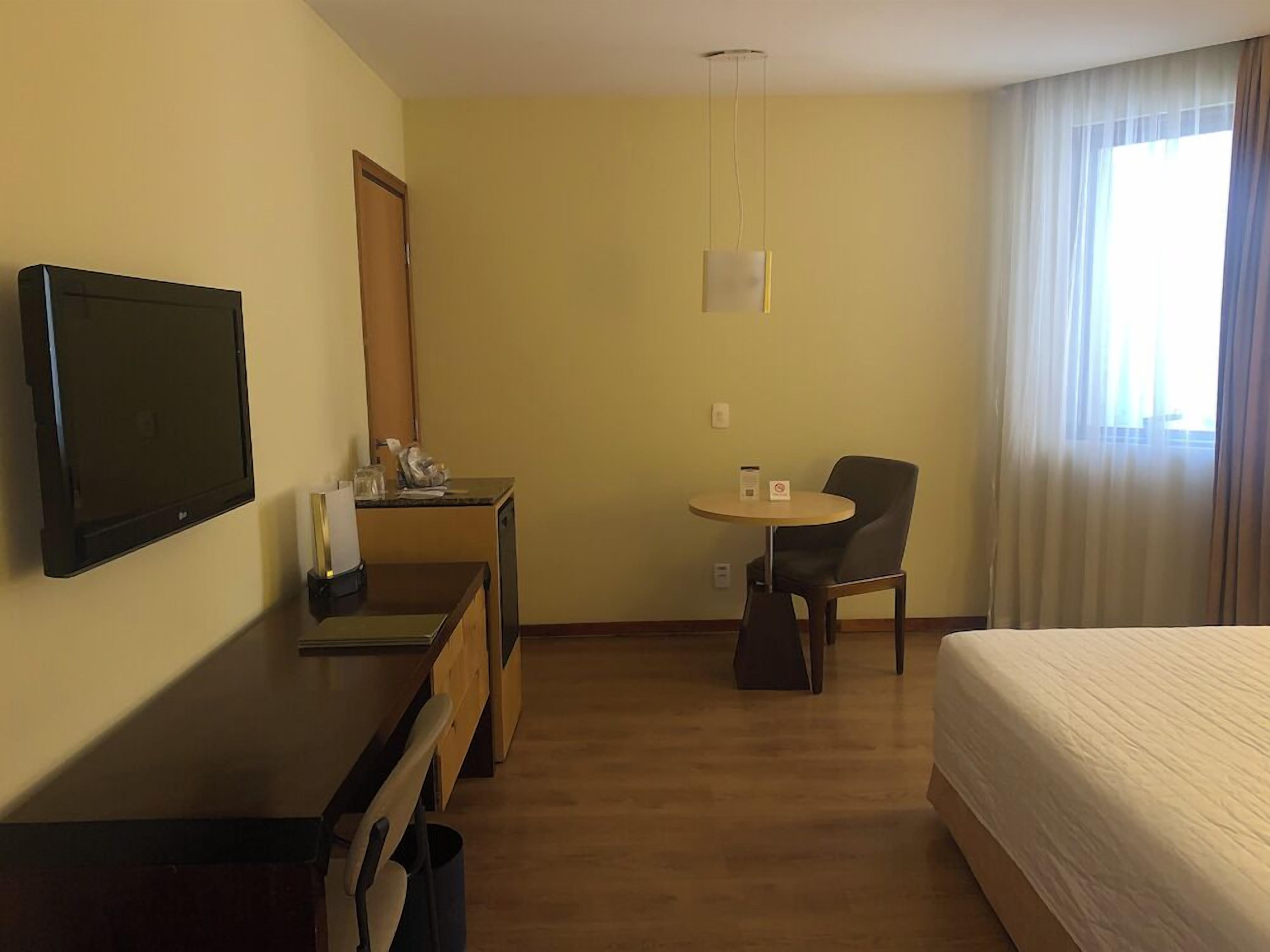 A hotel room with a bed, a desk with a television, a chair, a small table, and a window with curtains.