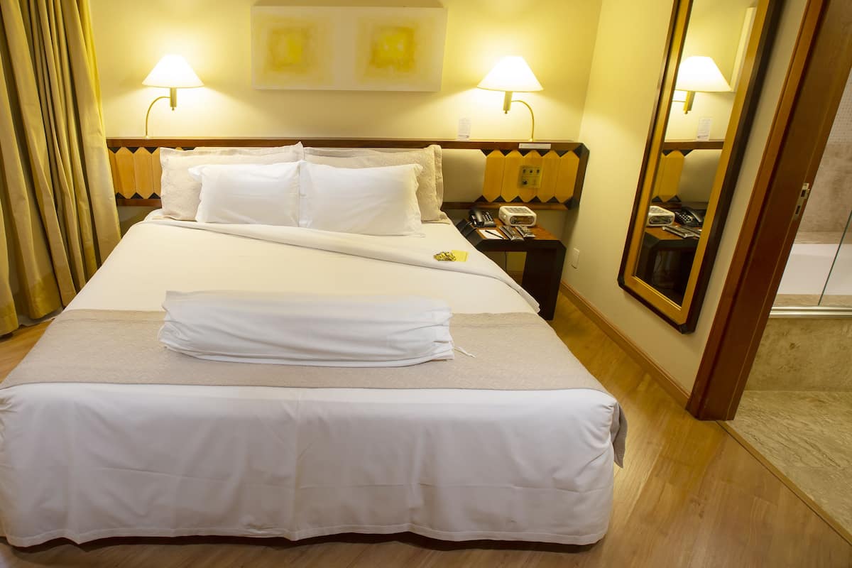 A hotel room with a neatly made bed, two bedside lamps, a bedside table with a phone and a lamp, a mirror, and a small wooden bench.