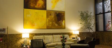 Un hall con un sofá, dos sillones, una mesa de centro y un jarrón con flores. Se observan pinturas abstractas en la pared y un ventanal con persianas.