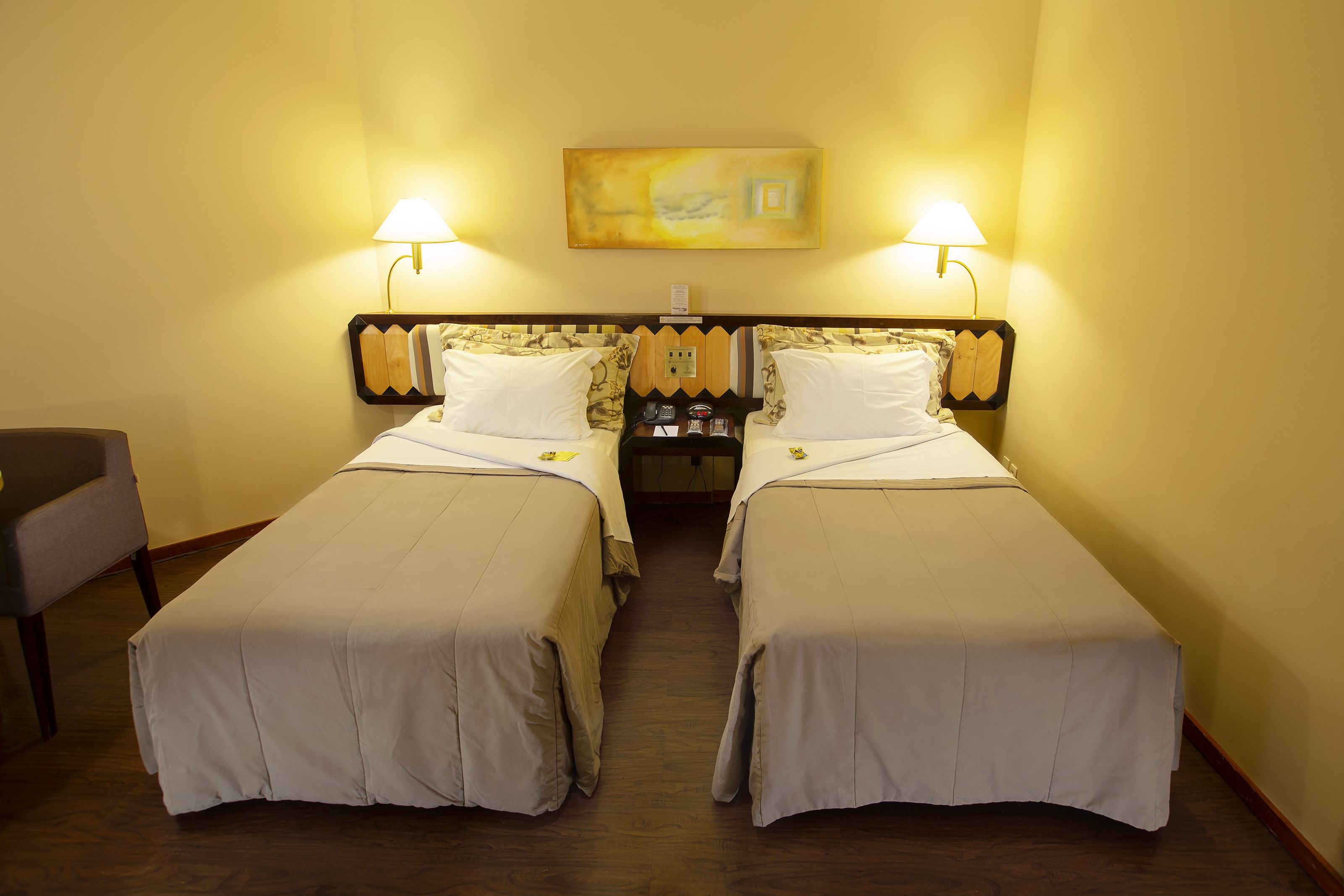 Una habitación de hotel con dos camas individuales, una mesita de noche con lámpara, una silla y un cuadro en la pared.