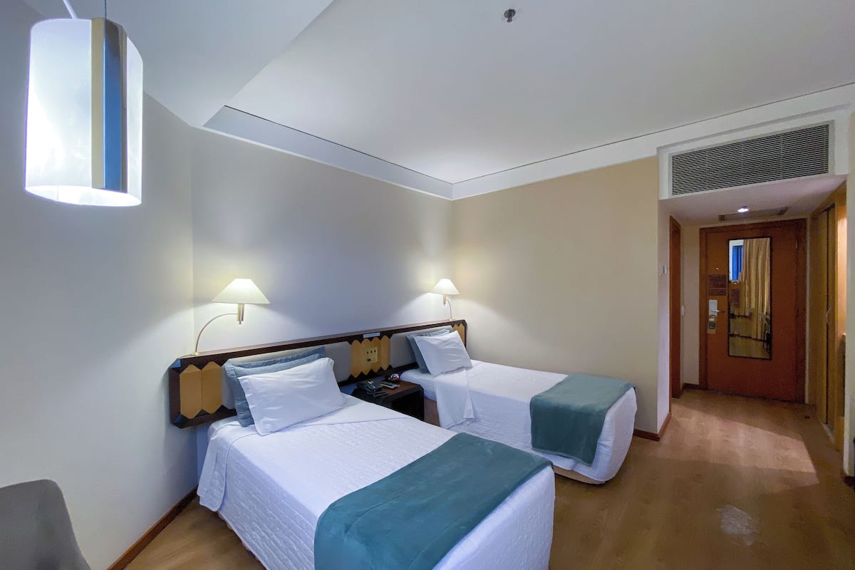 A hotel room with two beds, a desk, and a chair. There are two wall-mounted lamps and a ceiling light.