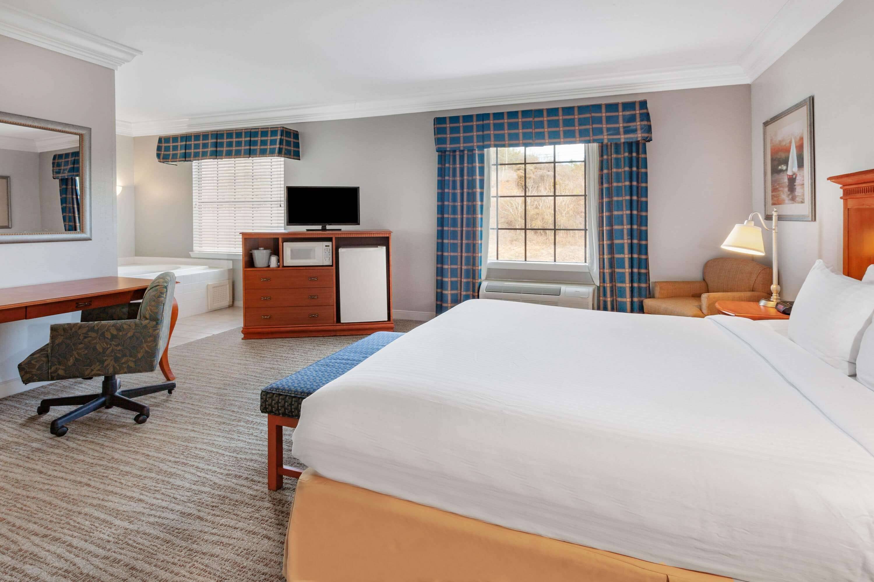 A hotel room with a large bed, a desk with a chair, a television, a dresser, and a window with curtains.
