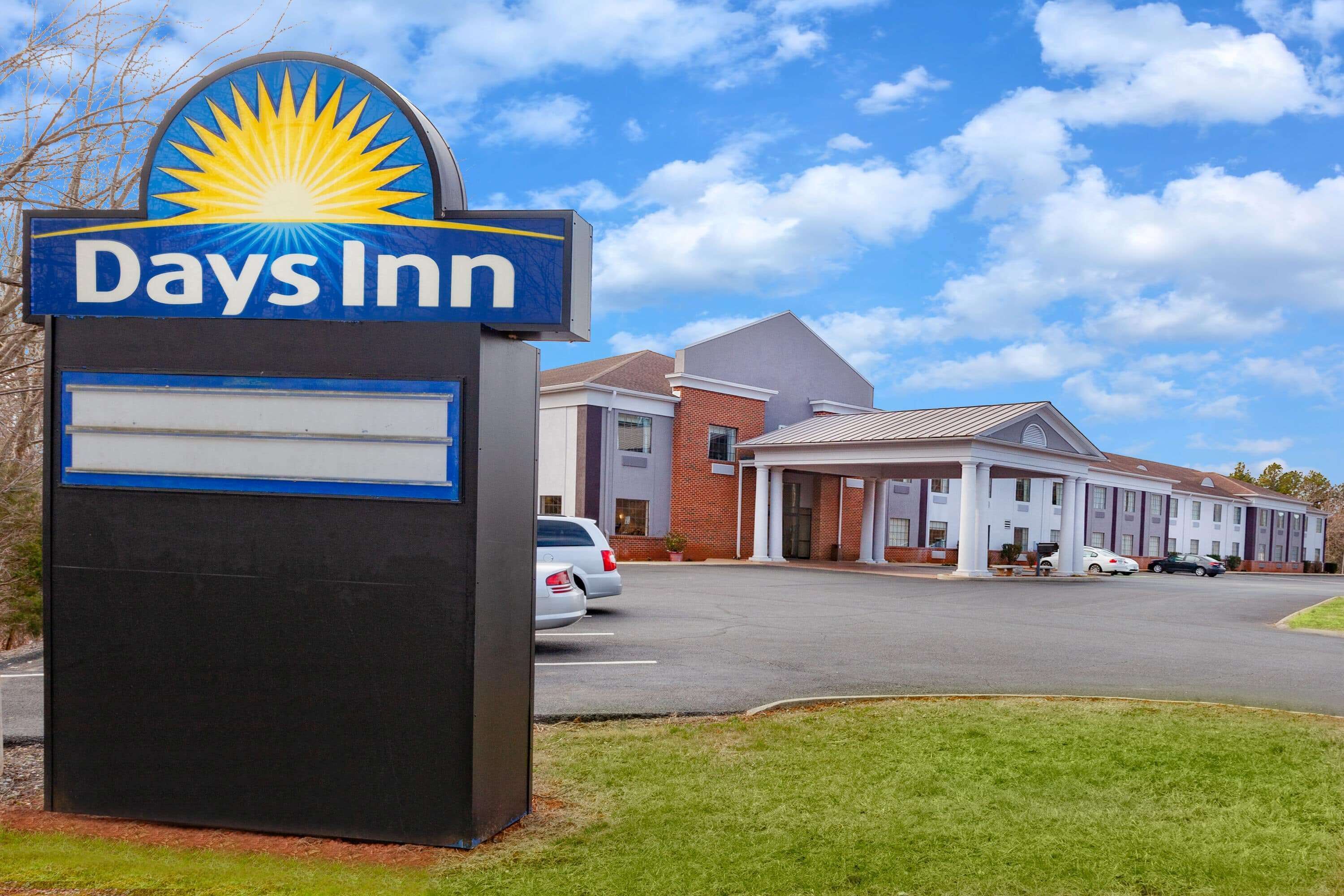 Señal de Days Inn con un emblema de sol, edificio de ladrillo con área de estacionamiento cubierta y un cielo azul despejado.
