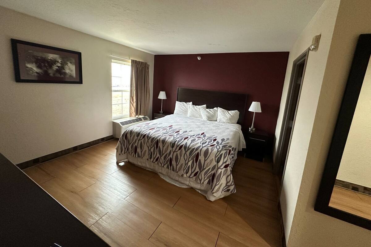 A hotel room with a bed, a nightstand, a lamp, a window with curtains, and a framed picture on the wall.