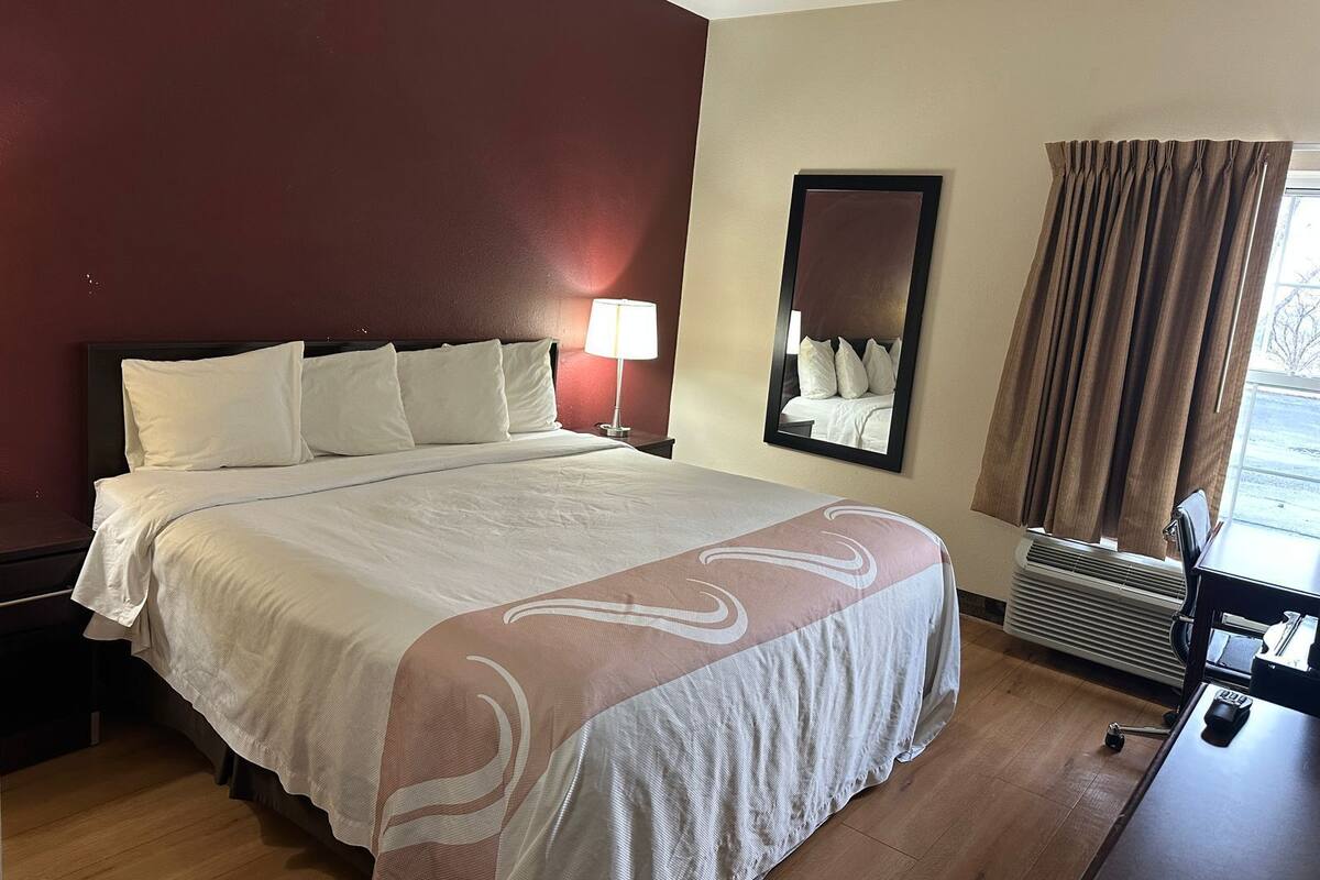 A hotel room with a bed, a nightstand, a lamp, a mirror, a window with curtains, and a heater.