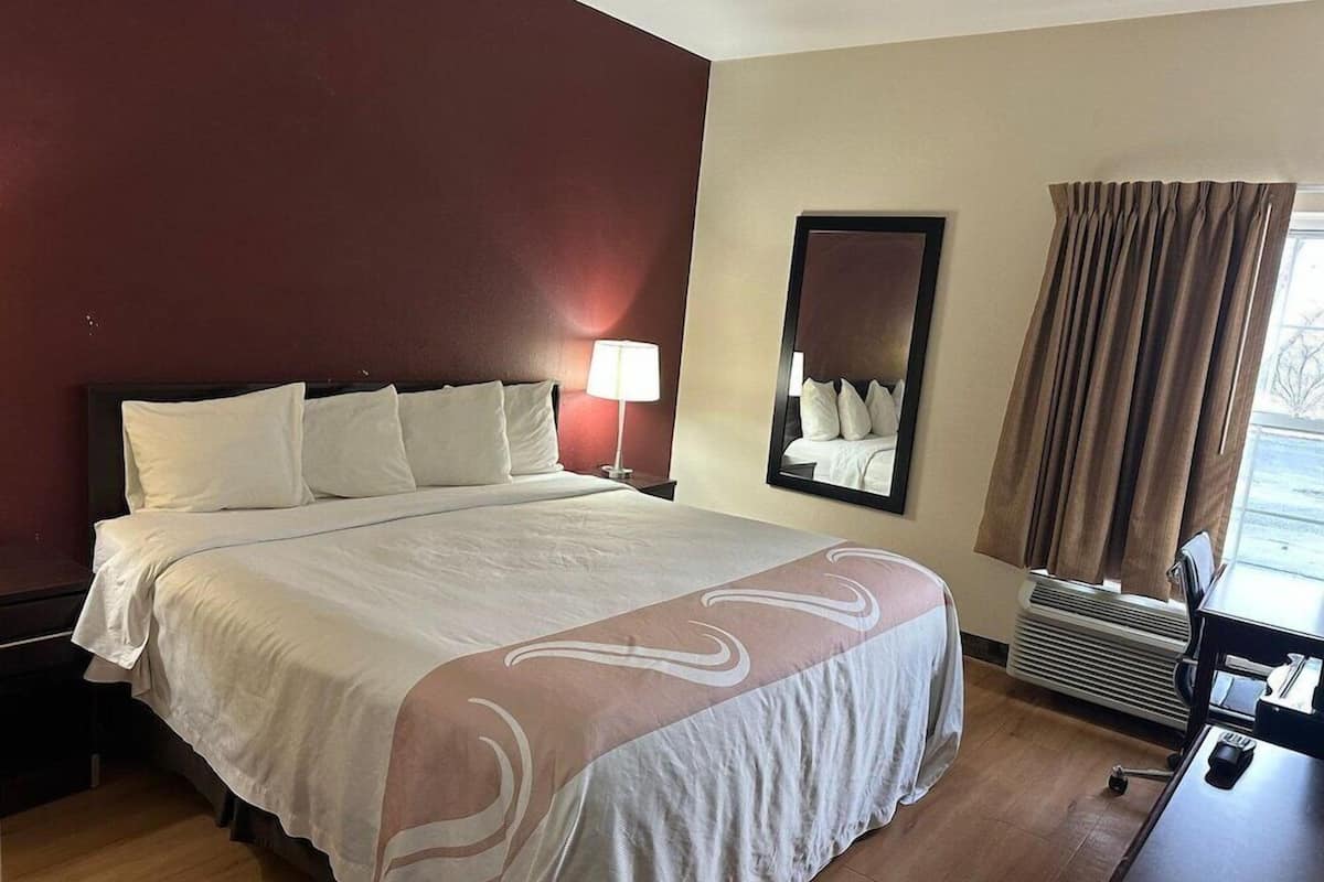 A hotel room with a large bed, a nightstand, a lamp, a mirror, a window with curtains, and a desk with a chair.