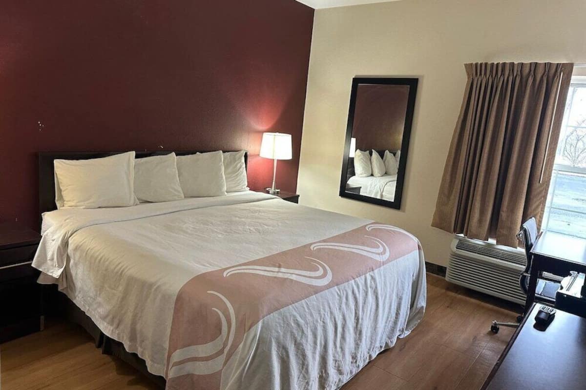 A hotel room with a large bed, a nightstand, a lamp, a mirror, a window with curtains, and a desk with a chair.