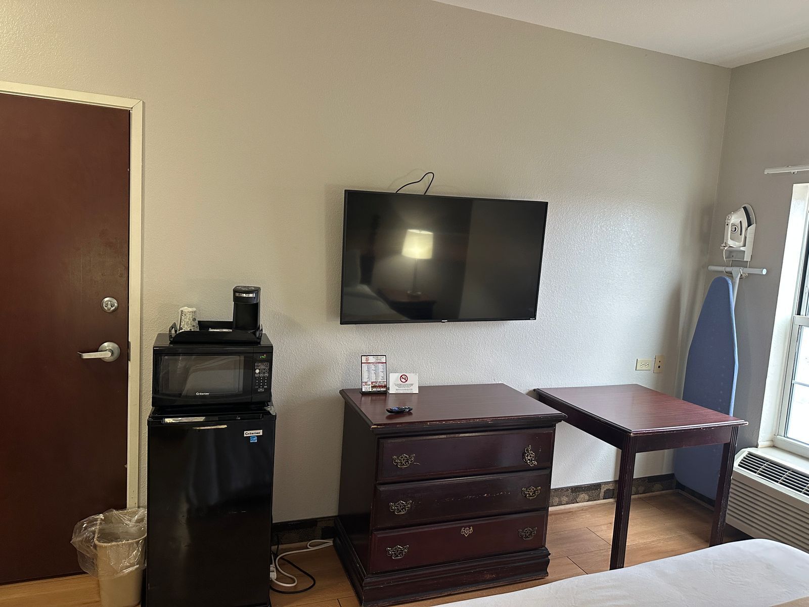 A hotel room with a television, a microwave, a dresser, a table, and a window with an ironing board.
