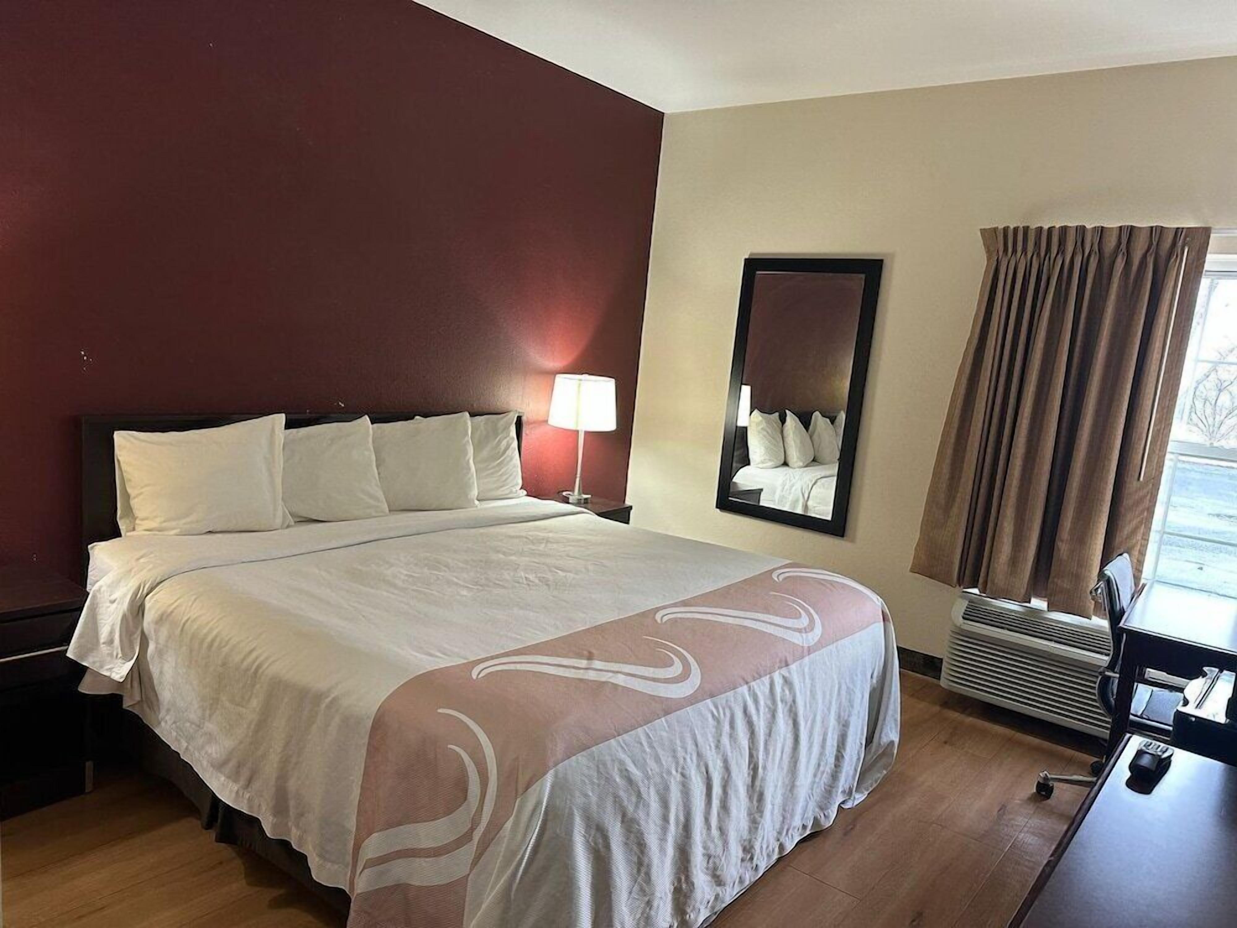 A hotel room with a large bed, a nightstand, a lamp, a mirror, a window with curtains, and a desk with a chair.
