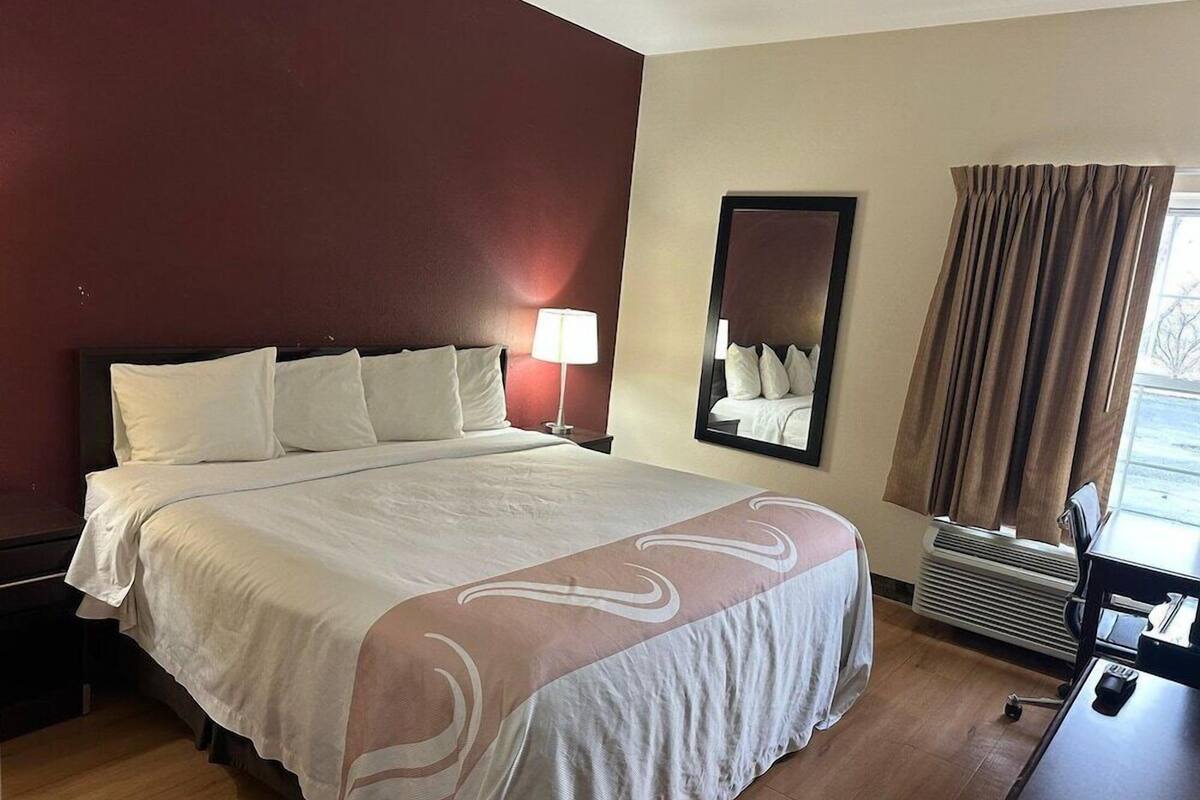 A hotel room with a large bed, a nightstand, a lamp, a mirror, a window with curtains, and a desk with a chair.