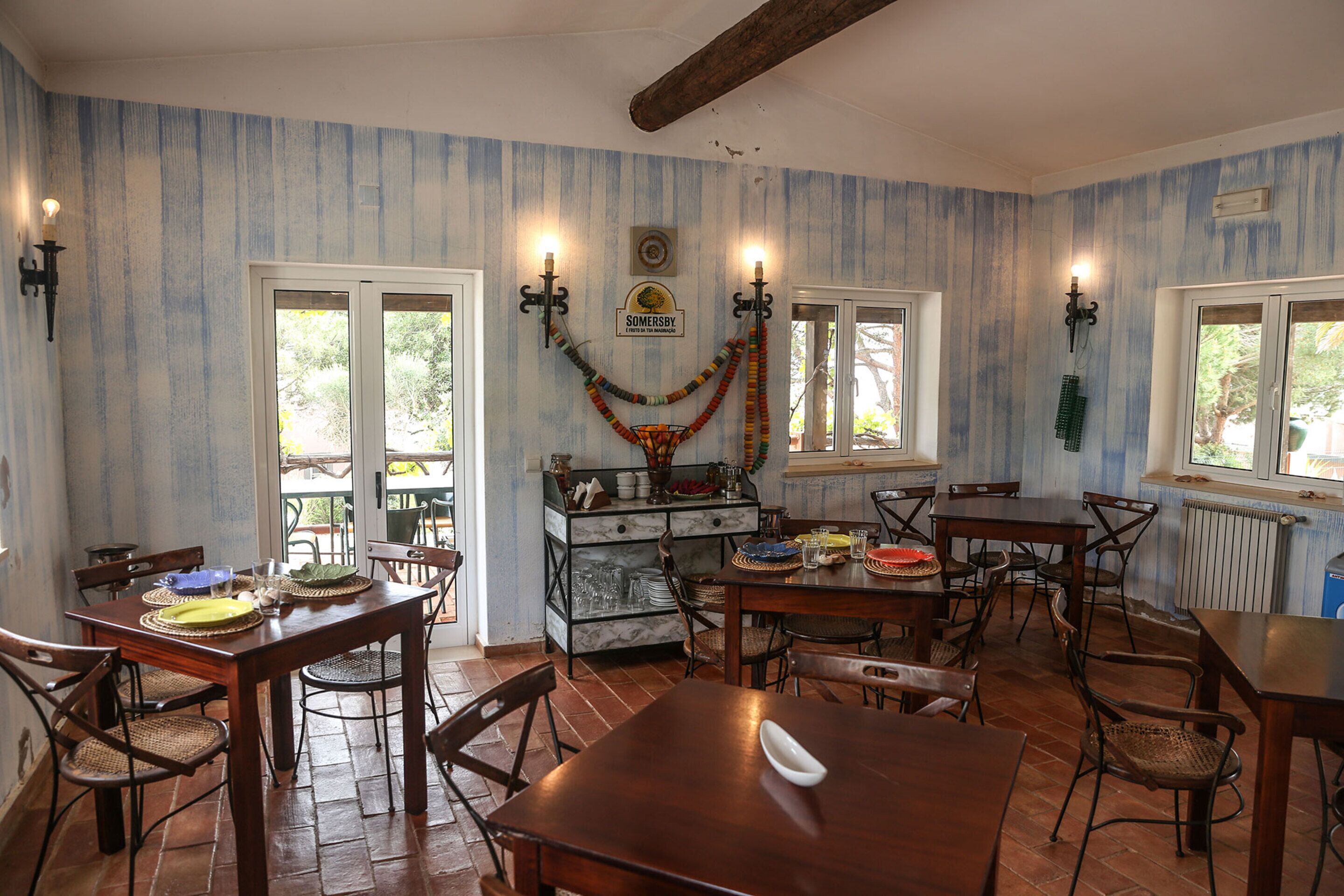 A dining area with wooden tables and chairs, a kitchen in the background, and a decorative garland on the wall.