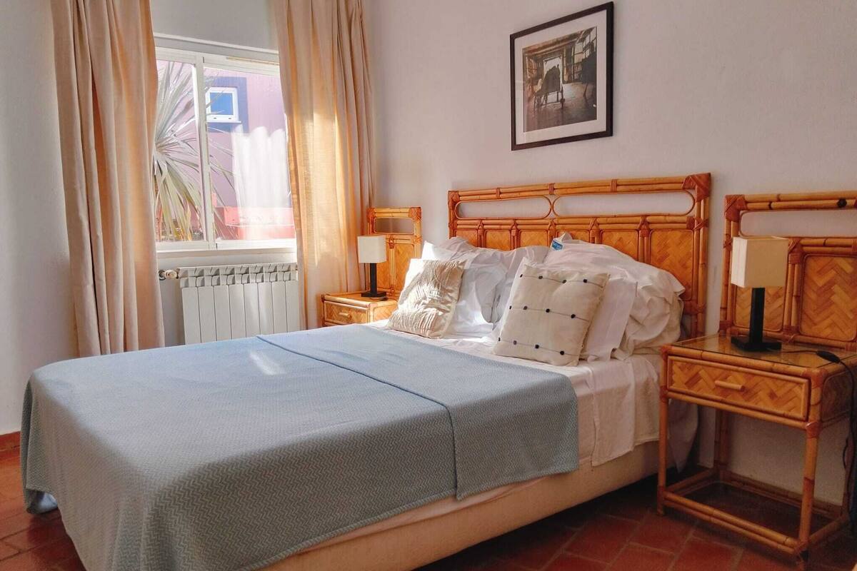 A neatly made bed with a blue bedspread, two bedside tables with lamps, a framed picture on the wall, and a window with curtains.