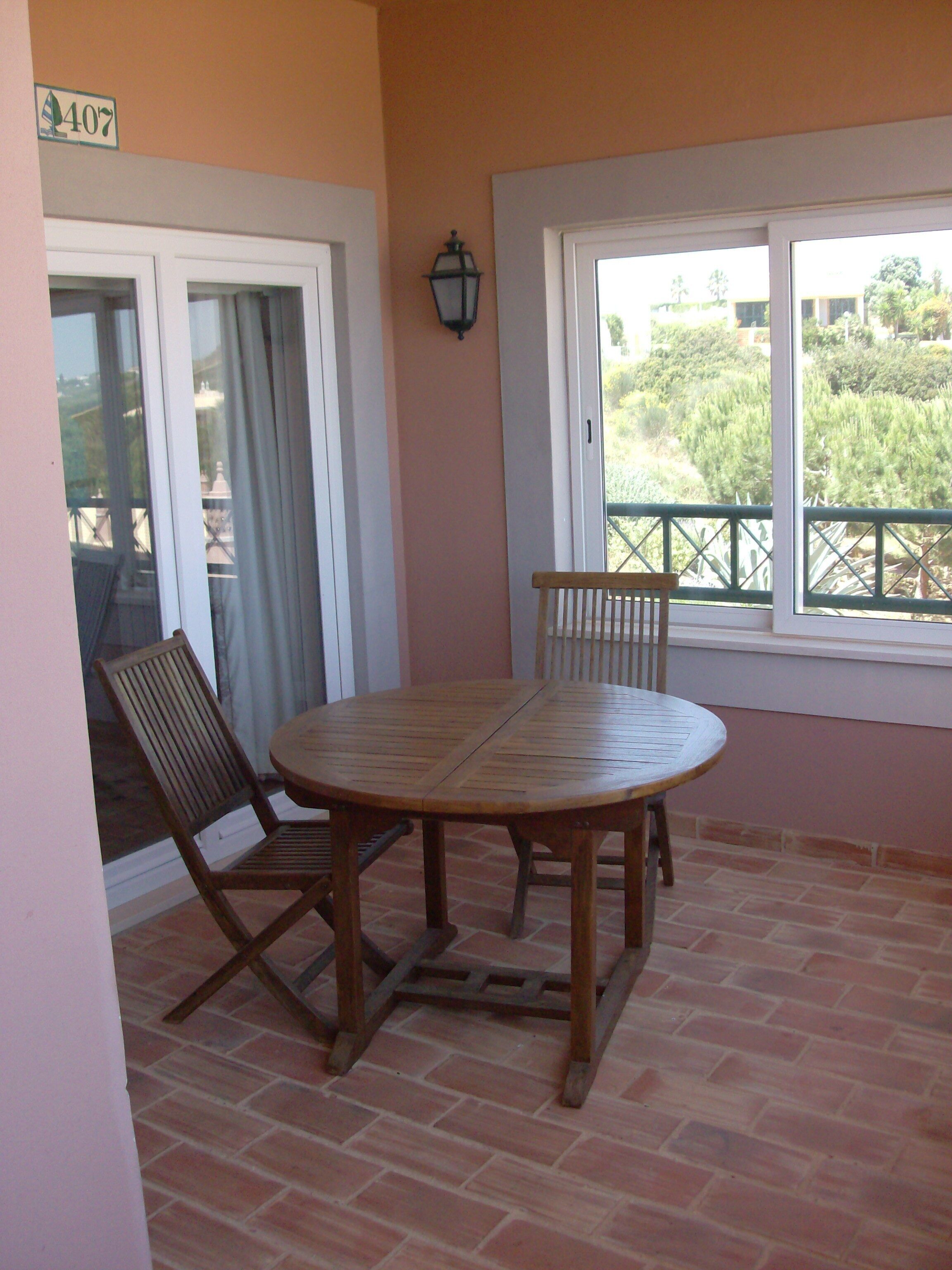 Un patio con una mesa y sillas de madera redonda, suelo de ladrillo y vista a través de una ventana.
