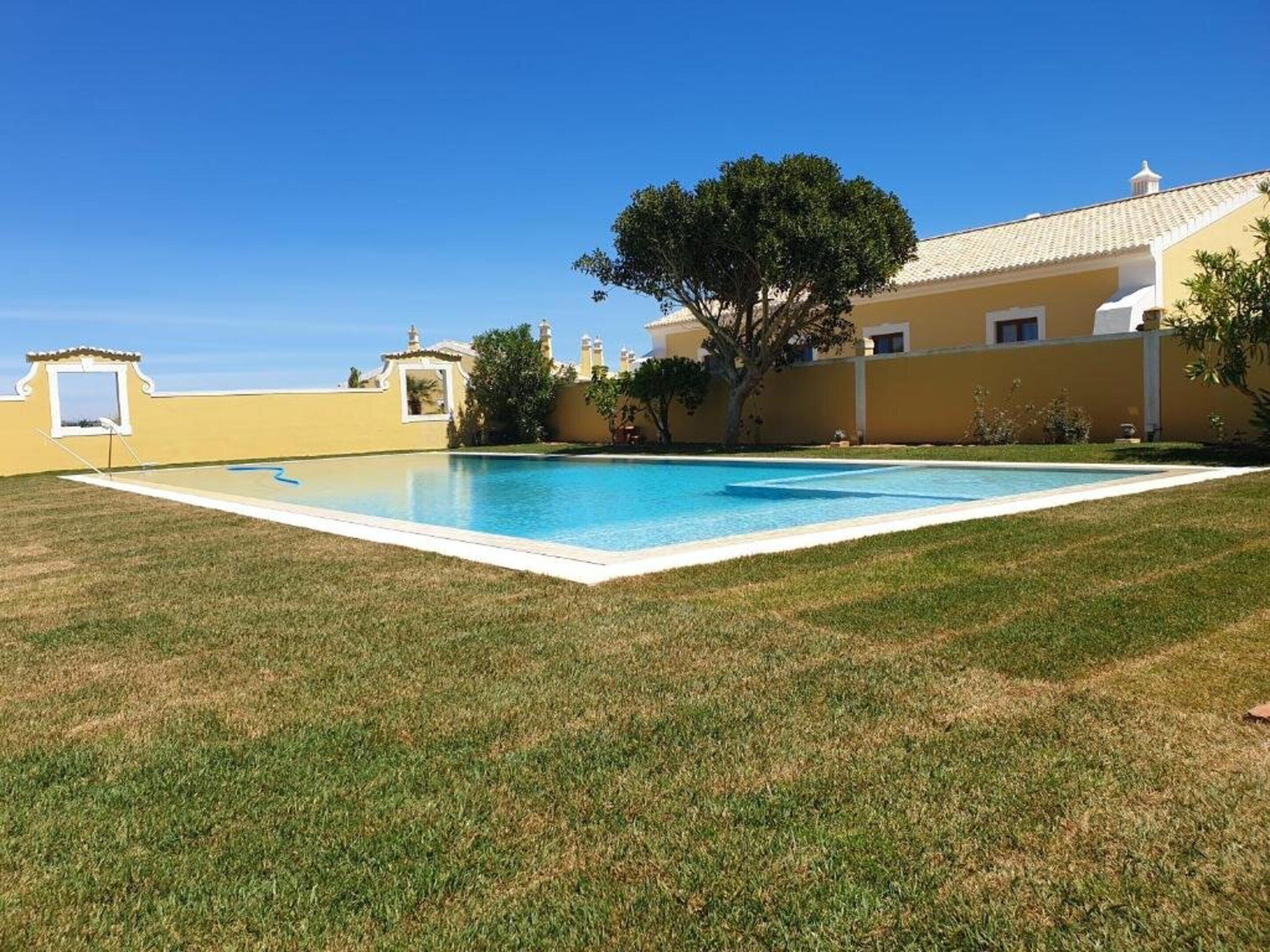A rectangular swimming pool with a white border, surrounded by a well-maintained lawn and a building with a yellow facade and white trim.