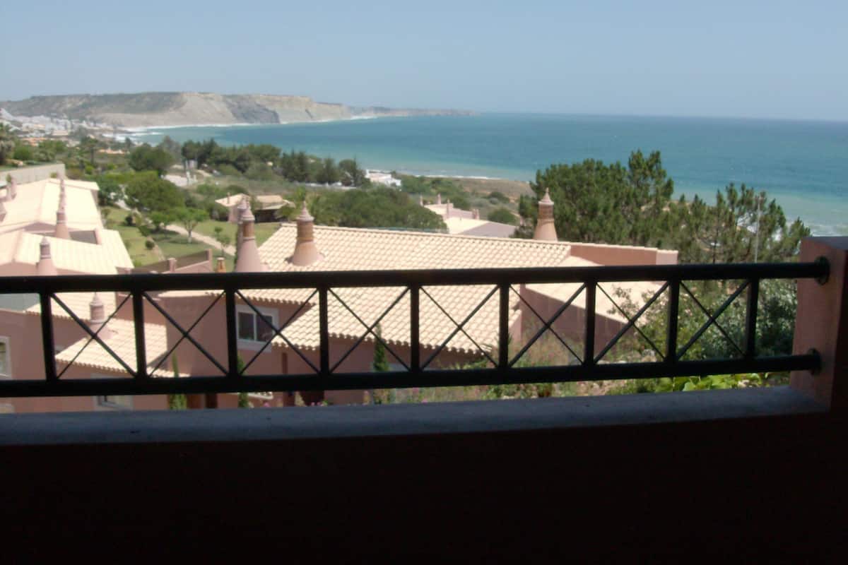A coastal view from a balcony with a railing, overlooking a beach and a distant coastline.